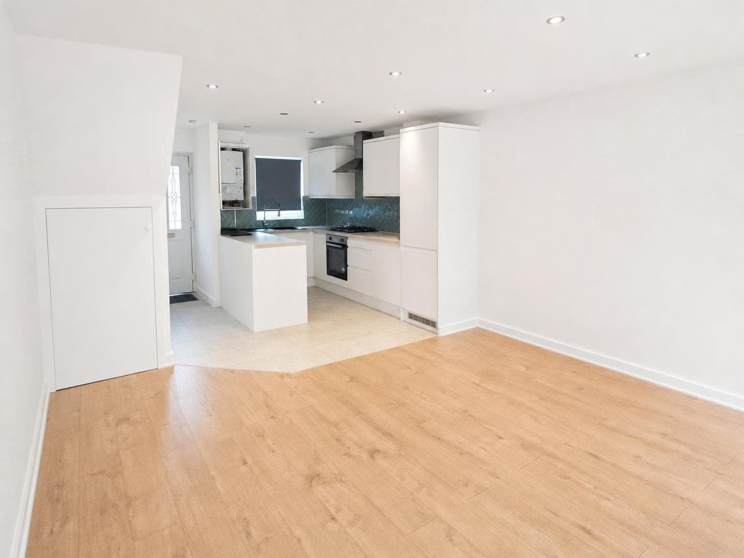 Open-plan kitchen and living room with white-painted walls, ceiling and skirting boards, pale wooden flooring, white kitchen cabinets and dark backsplash.