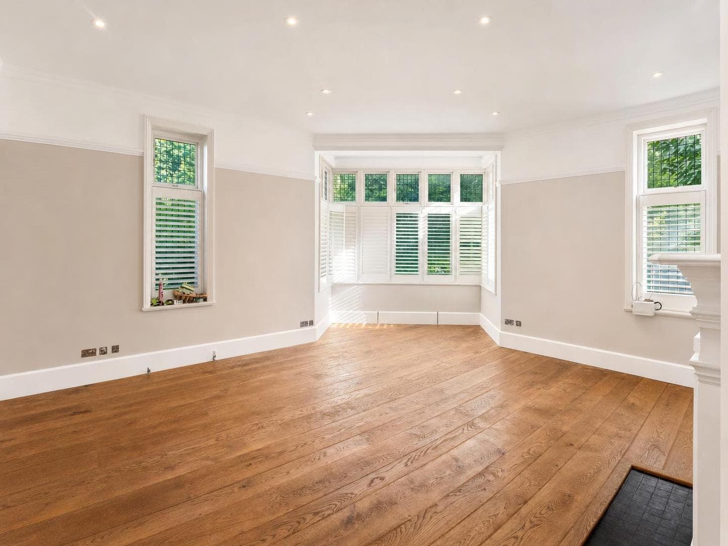 Reception room with neutral painted walls, white skirting boards and architraves, wooden flooring and multiple windows with shutters