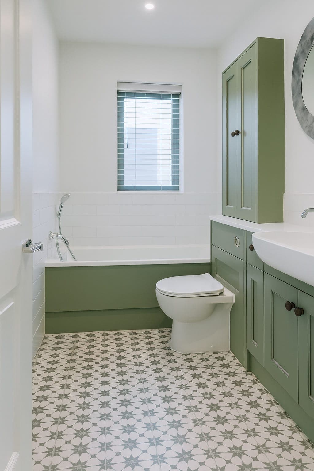 Bathroom with sage green lower wall and matching vanity and tall cabinet. Upper walls and ceiling white. Patterned floor tiles and green-framed window.