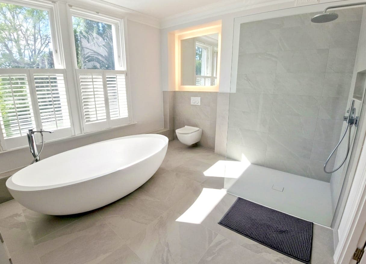 Bathroom with white painted walls and ceiling, freestanding white bath, walk-in shower, wall-mounted basin with lit mirror, and window with white shutters.
