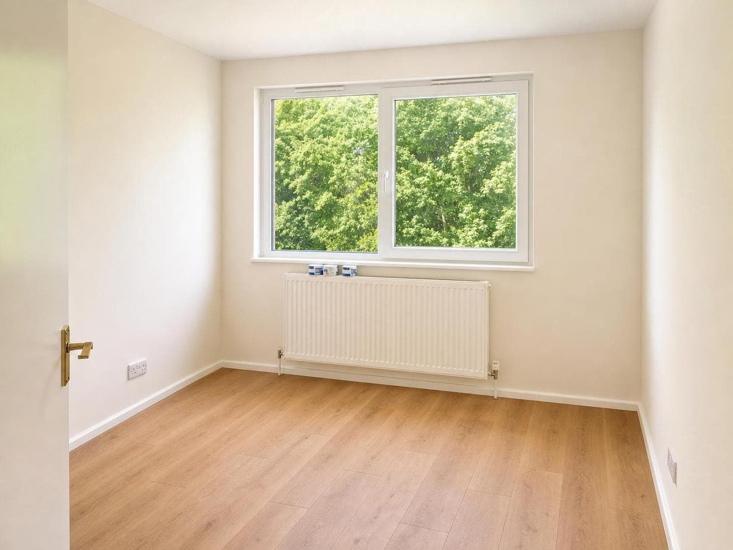 Bedroom with pale walls, white ceiling, cream radiator and white skirting boards; large window overlooks green trees.